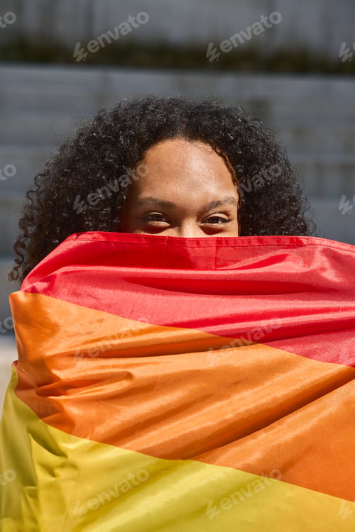 Preview: Young woman hiding behind progress pride flag looking at camera