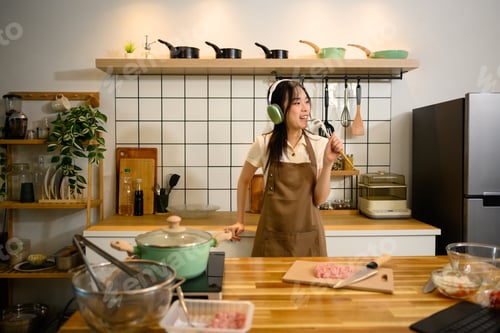 Preview: Cheerful woman wearing head phone singing while cooking in a stylish kitchen