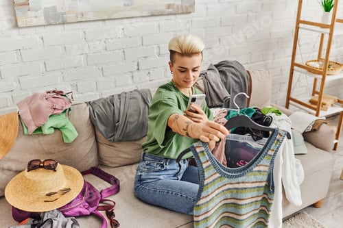 Preview: clothing swapping, tattooed woman taking photo of knitted tank top on smartphone on couch at home
