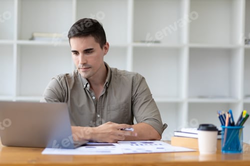 Preview: Handsome young businessman in office working on laptop and financial documents.