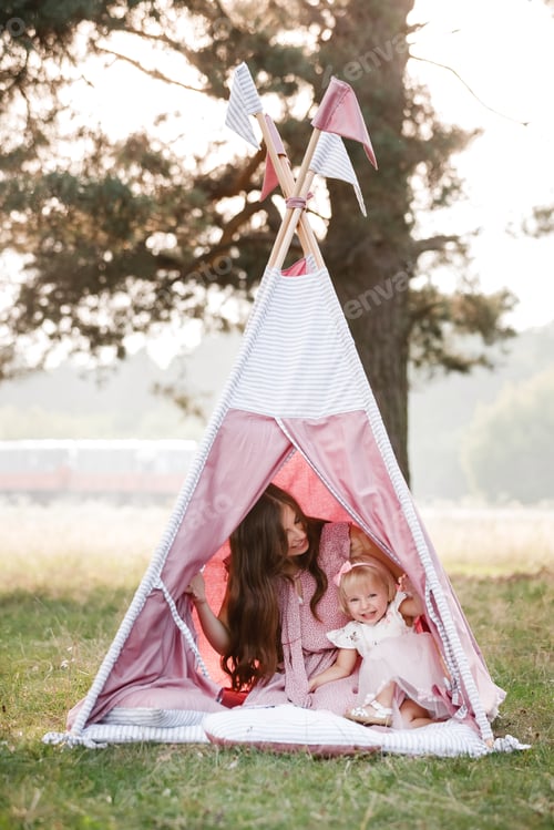 Preview: Beautiful mother with her little daughter sitting near wigwam in the park. Spending time together,