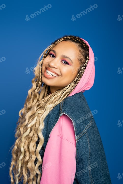 Preview: joyful and pretty african american woman with hood on head smiling on blue background, positive