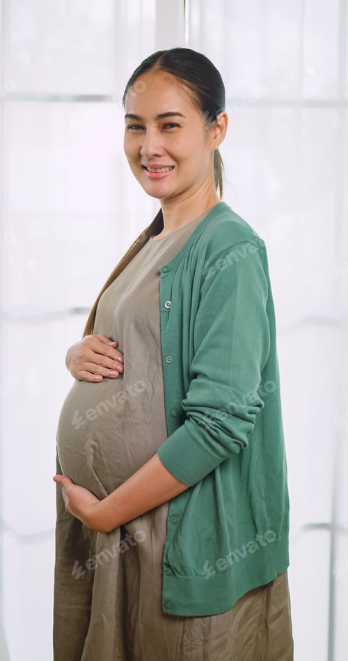 Preview: Asian pregnant woman lovingly touching her unborn child while standing by the window of the house.