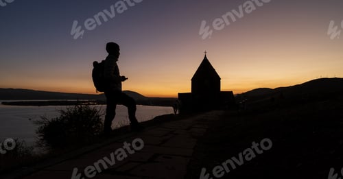 Preview: Silhouette of a traveler walking toward a church at sunset by the lake.