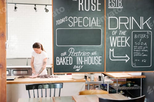 Preview: Woman wearing a white apron standing at a work counter in a bakery, menu on a blackboard.