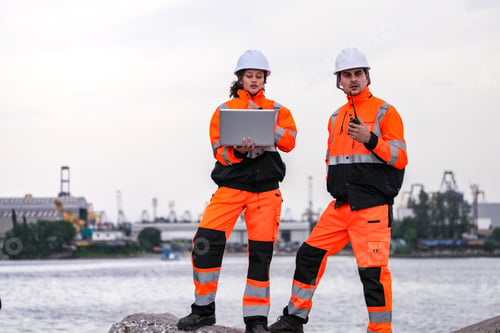 Preview: Two engineers wearing gear review plans on a laptop at a shipyard,