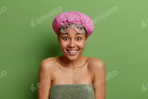 Preview: Positive dark skinned woman smiles broadly going to take shower wears bath hat waterproof goggles wr