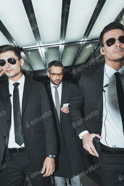 Preview: businessman looking at smartphone while standing with bodyguards in elevator