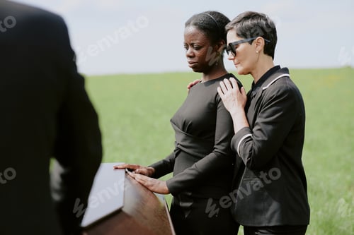 Preview: Caucasian Woman Comforting Black Woman Mourning Beside Coffin Outdoors