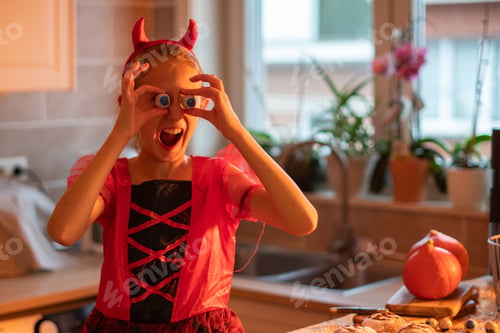 Preview: Caucasian girl in devil costume showing scary faces while decorating house with Halloween decor, kid