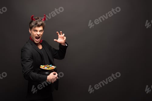 Preview: sexy scary man in halloween devil costume holding pot with candies on black