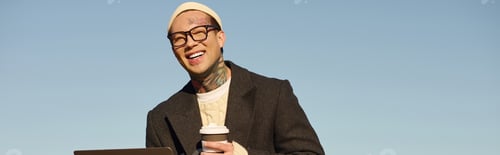 Preview: Handsome stylish man with tattoos enjoying coffee while working outdoors on a sunny day