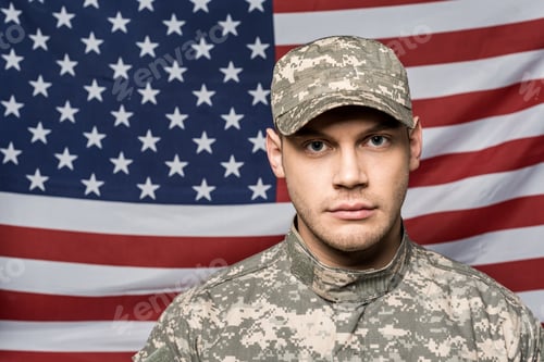 Preview: handsome soldier in military uniform and cap looking at camera near flag of america