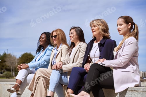 Preview: Group of businesswomen looking away while sitting on a bench outdoors.