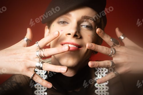 Preview: smiling queer person in black beret and silver rings posing with hands near face on dark red