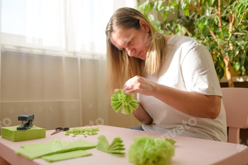 Preview: A woman in the rays of the sun makes paper flowers from green napkins at a table by the window.
