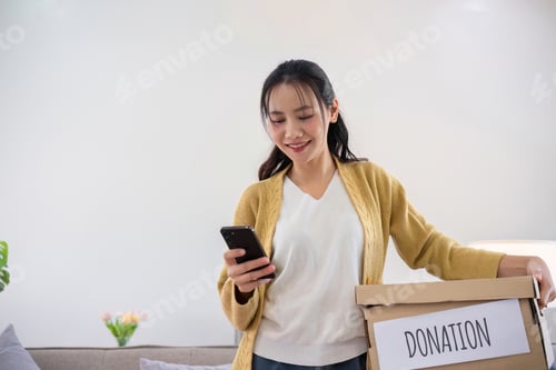 Preview: A cute Asian woman stands holding a box, sorting unwanted clothes for donation.