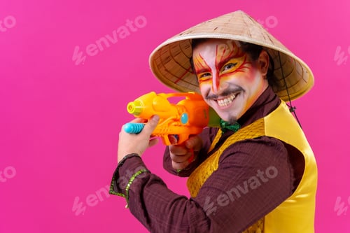 Preview: Clown with white facial makeup on a pink background, smiling with toy guns