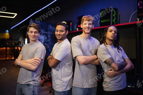 Preview: Smiling multiethnic gamer team in grey t-shirts crossing arms in gaming club