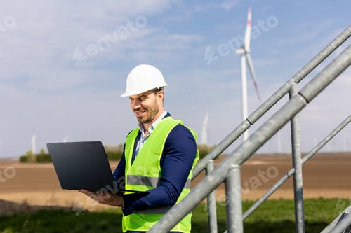 Preview: Engineer in High-Visibility Vest Analyzing Data on Laptop at Wind Farm During Daytime