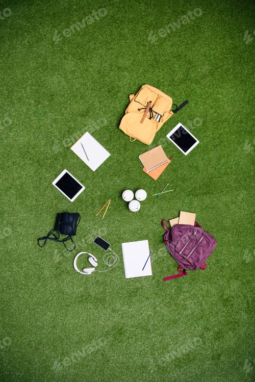 Preview: top view of arranged digital devices, backpacks and notebooks on green lawn