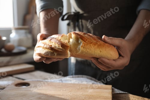 Preview: Man breaking loaf of fresh bread at wooden table indoors, closeup