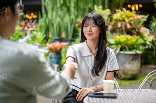 Preview: Glasses man and pretty woman doing handshake while sitting together at round table in cafe's garden.