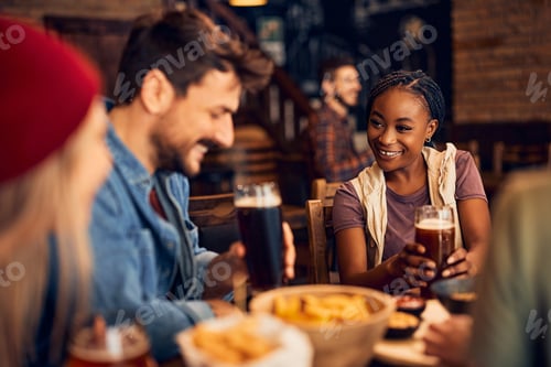 Preview: Happy black woman talking to her friends while drinking beer in pub.