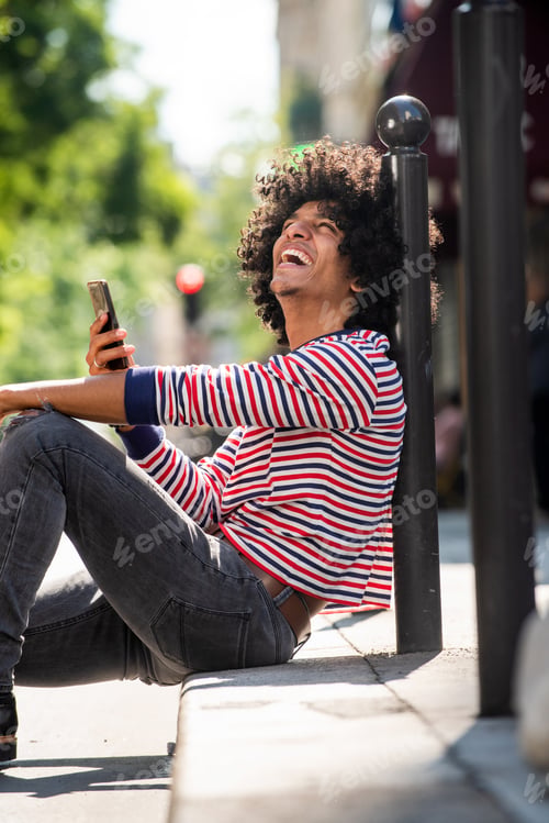 Preview: happy young man with afro hair holding mobile phone while sitting on city street