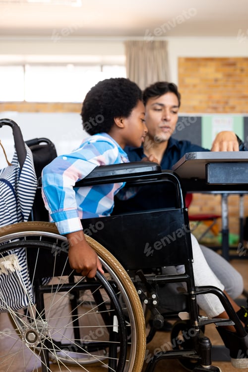 Preview: Diverse boy in wheelchair and male teacher in classroom at elementary school