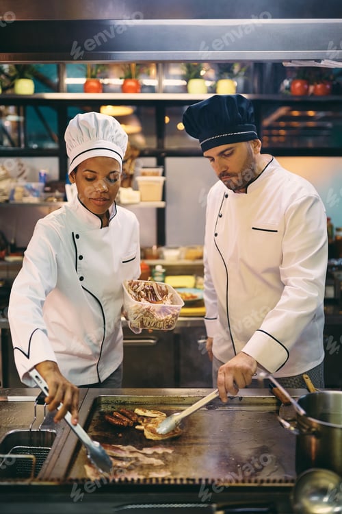 Preview: Mid adult chef and his black female colleague preparing food together at restaurant kitchen.