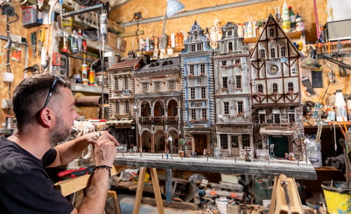 Preview: Modeller man working at scale model of miniature building in his workshop full of tools