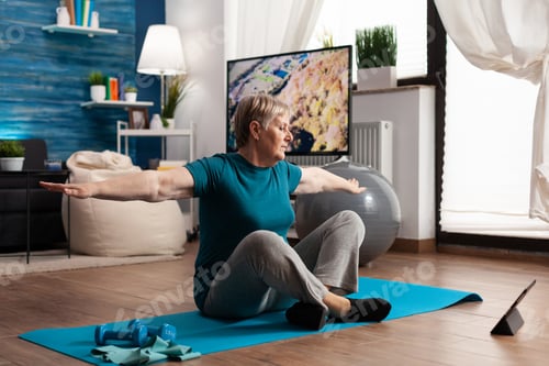 Preview: Retired senior woman eatching fitness tutorial on laptop sitting on yoga mat streching arm