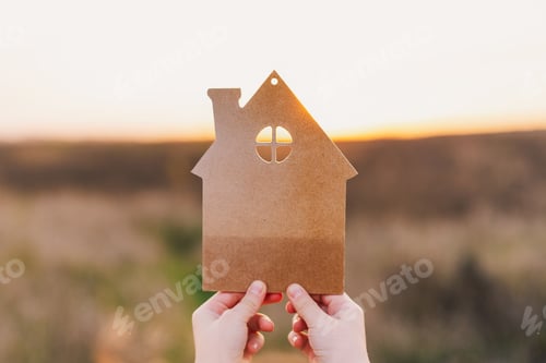 Preview: Man's hand holds a wooden house on the background of blurred buildings