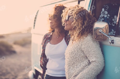 Preview: Two adult young woman enjoying time and leisure together against a classic blue van in travel
