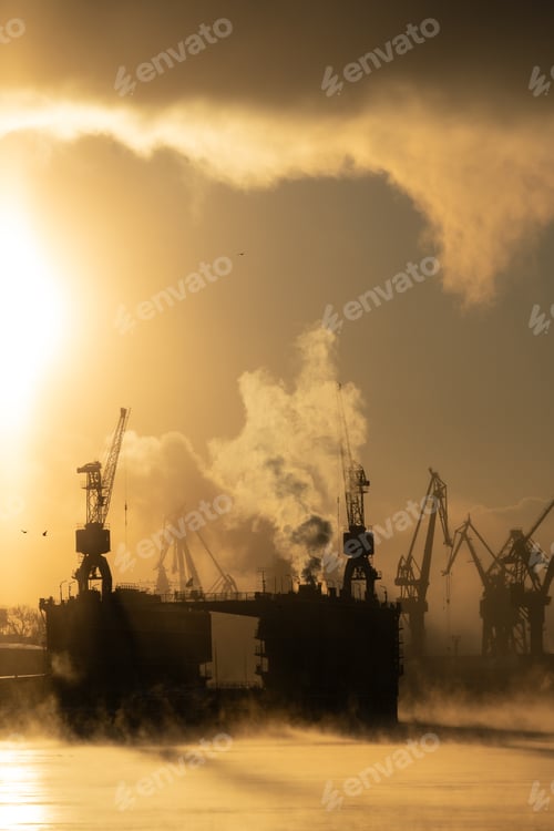 Preview: Cranes of shipyard in frosty winter day, steam over river, smooth surface of river at sunset.
