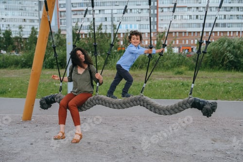 Preview: Active curly little boy riding on swing at playground with mom. Childhood, family leisure. Happy