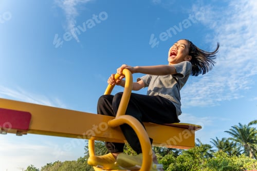 Preview: A happy young Asian girl on a yellow seesaw at a playground.