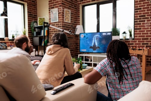 Preview: Multiethnic group of friends sitting on sofa at home while playing space videogame on gaming console