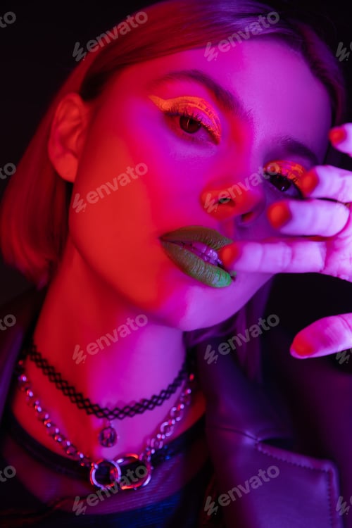 Preview: portrait of young woman with bright neon makeup holding hand near face in purple light on black