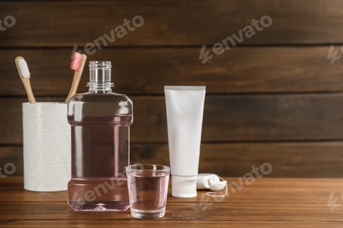 Preview: Bottle and glass with mouthwash near other oral hygiene products on wooden table, space for text