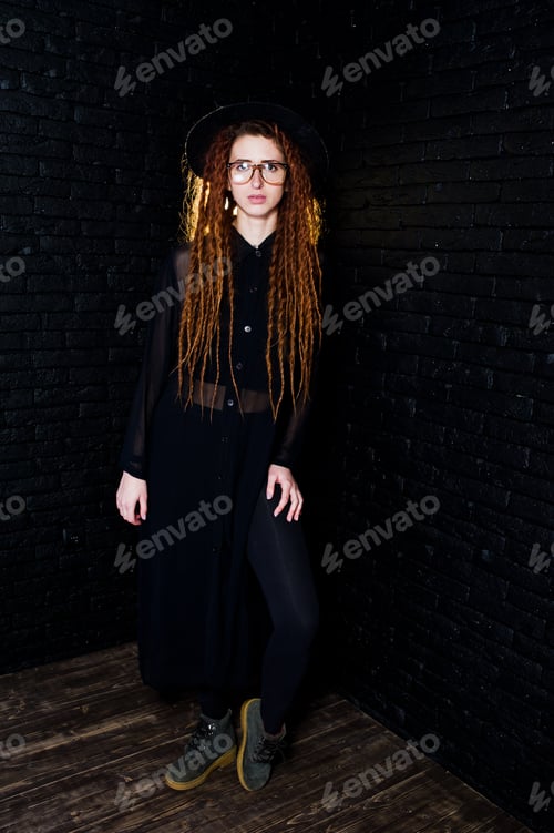 Preview: Studio shoot of girl in black with dreads, at glasses and hat on brick background.