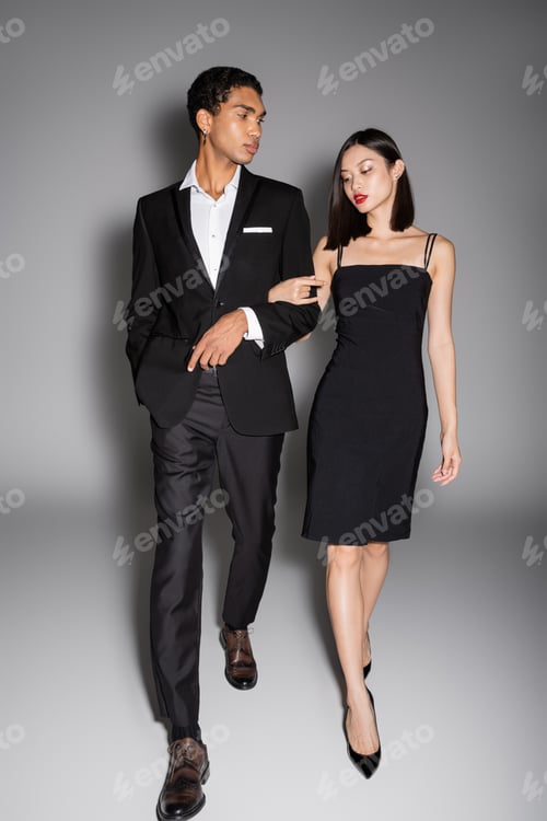 Preview: full length of multiethnic couple in black elegant attire walking on grey background
