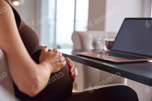 Preview: Close Up Of Pregnant Woman Working From Home On Laptop Sitting At Table Holding Stomach