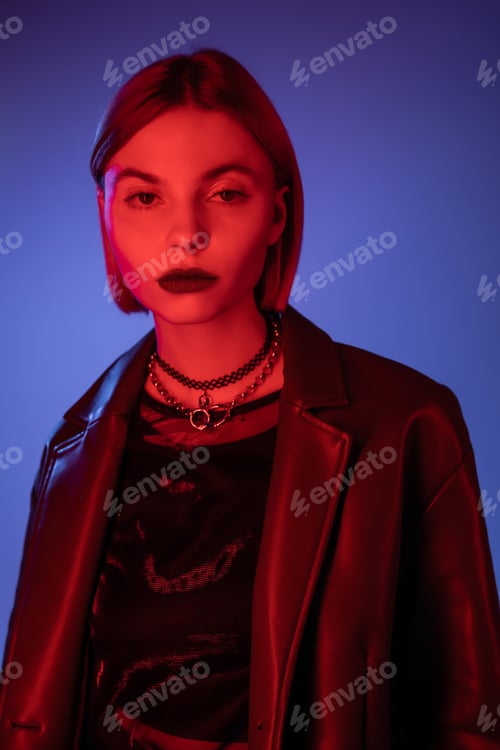 Preview: young woman in leather jacket looking at camera in red neon light on blue background