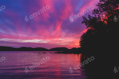 Preview: Beautiful purple and pink sunset over a lake with mountains in background and silhouette of trees
