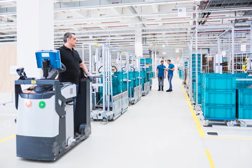 Preview: Two men with tablet talking in industrial hall with man on tugger train