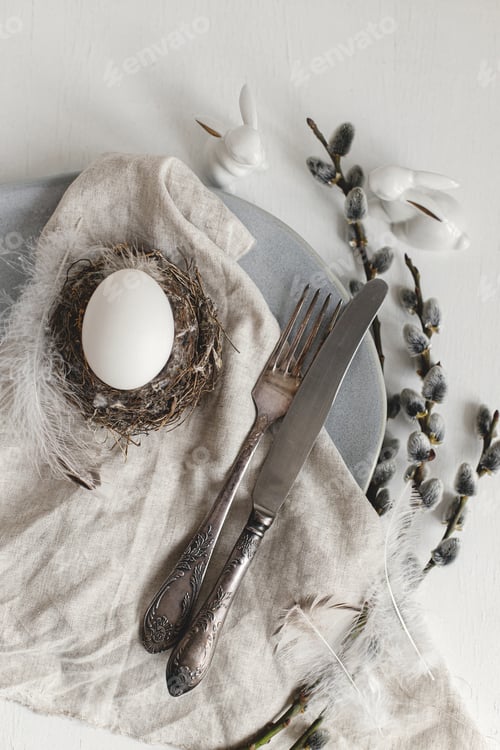 Preview: Stylish Easter table setting flat lay. Easter egg in nest, willow branches, feathers on modern plate