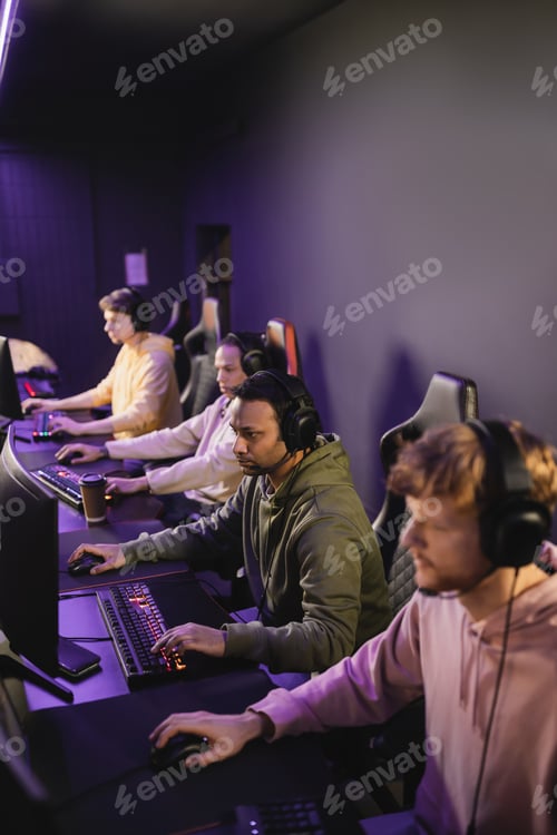 Preview: Indian gamer in headphones with microphone playing video game on computer near team in cyber club