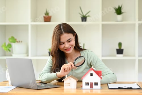 Preview: Smiling young female appraiser looking at house model through magnifying glass.